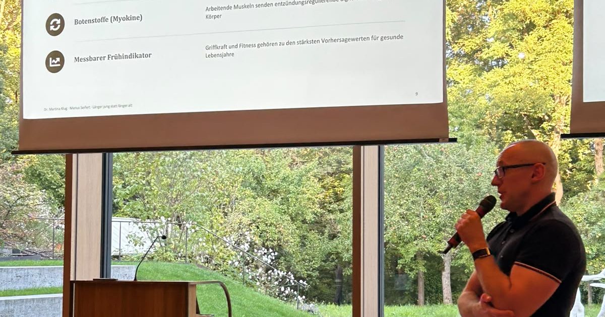 Marius Seifert spricht mit Mikrofon vor großen Fenstern, links das Rednerpult, darüber eine Folie mit den Punkten Botenstoffe (Myokine) und Messbarer Frühindikator